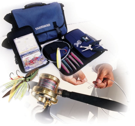 Hands tying a fishing knot with a rod and reel, surrounded by Shimano tackle, lures, and fishing tools